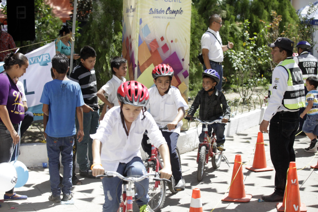 Kinder fahren mit Fahrrädern eine Straße entlang, an der Verkehrshütchen stehen, einige tragen Helme, andere stehen daneben, mit einem Banner, Bäumen und Gebäuden im Hintergrund.