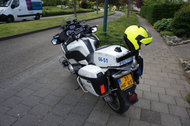 Ein Polizeimotorrad am Straßenrand geparkt mit einem Helm darauf, mit Bäumen, Pfählen, Gebäuden, Pflanzen, Gras, Steinen und anderen Fahrzeugen im Hintergrund.