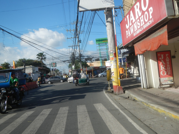 Eine belebte Stadtstraße mit Fahrzeugen, Fußgängern, Strommasten, Gebäuden, Bäumen und einem bewölkten Himmel, mit einem Geschäft auf der rechten Seite mit einer "Jaro Pfandleihe"-Schilder.