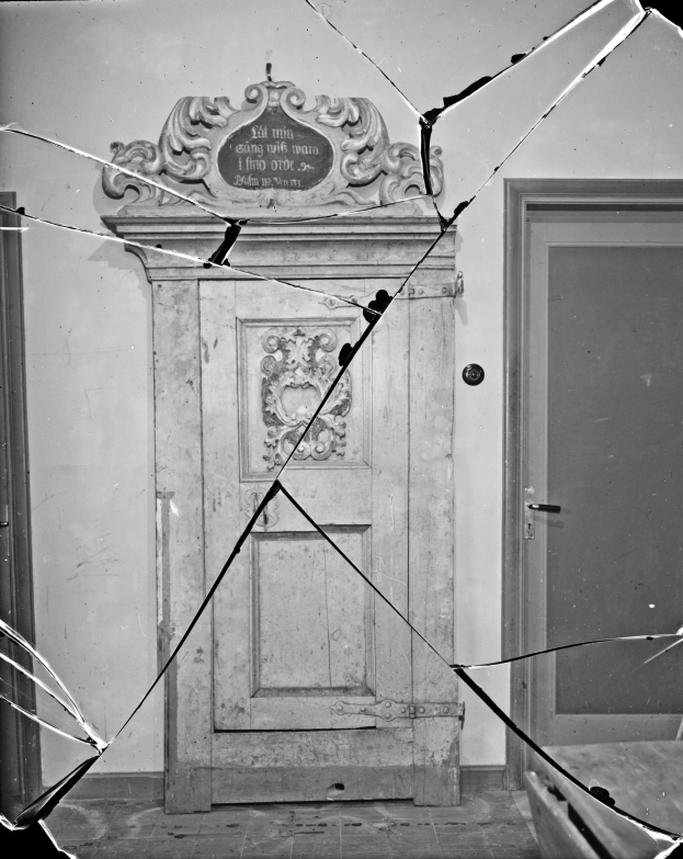 Schwarz-weiß-Foto einer zerbrochenen Tür in einem Raum mit Glasscherben im Hintergrund und einer Wand im Hintergrund.