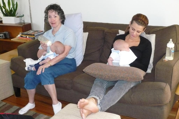 Zwei Frauen auf einer Couch sitzend, die Babys halten, mit Kissen auf der Couch, einer weißen Wand im Hintergrund und einem Schreibtisch mit Büchern und einer Zimmerpflanze links.