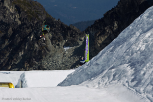 Ein Mann mit Skiern springt in die Luft, mit schneebedeckten Bergen und einer Flagge mit Text im Hintergrund, und ein anderer Mann steht vor der Flagge, mit Text in der unteren linken Ecke angezeigt.