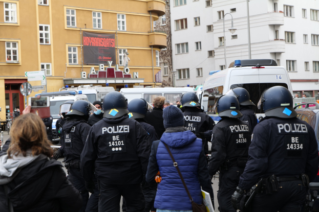 Eine Gruppe von Polizisten in Uniform steht vor einer Menge von Menschen mit Helmen und Jacken während einer Demonstration in Berlin, Deutschland, mit Fahrzeugen, Gebäuden, Laternen und einem Banner im Hintergrund.