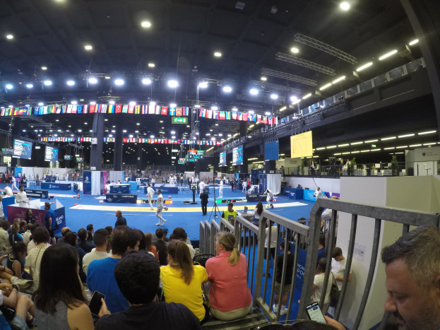 Eine große Menschenmenge sitzt und steht in einer Indoor-Sportarena, mit Bildschirmen, Fahnen und Deckenleuchten im Hintergrund.