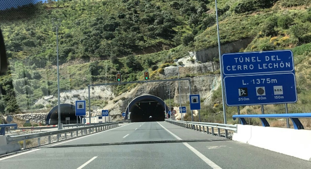 Auto fährt durch einen Tunnel neben einer Straße mit Geländern, Schildern, Pfosten, Ampeln, Bäumen, Felsen und einem klaren blauen Himmel.