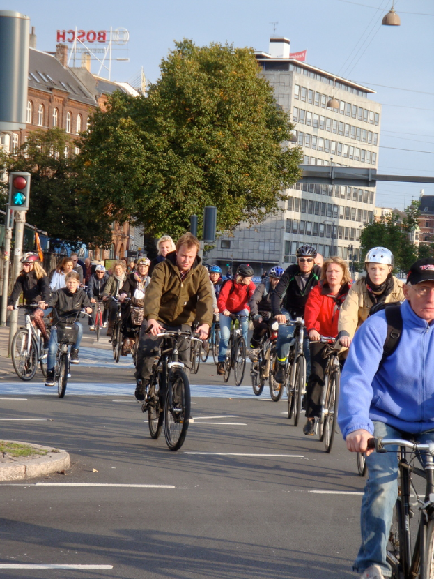 Menschen, die auf einer Straße mit Pfählen, Bäumen, Gebäuden und dem Himmel im Hintergrund radfahren.