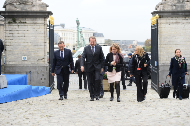 Eine Gruppe von Mitarbeitern geht in einen Bereich mit einem Tor, hinter dem sich Fahrzeuge und Gebäude befinden; rechts trägt eine Frau zwei Gepäckstücke.