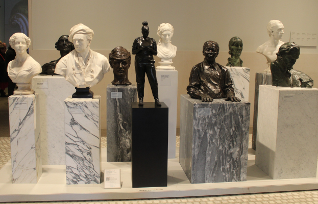 Eine Museumsausstellung von detaillierten, lebensechten Büsten-Skulpturen mit Besuchern im Hintergrund vor einer Wand.