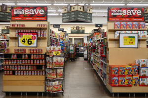 Ein Innenraum eines Lebensmittelladens mit verschiedenen Lebensmitteln auf Regalen und dem Boden, Preistafeln, Deckenleuchten und einer Person im Hintergrund.
