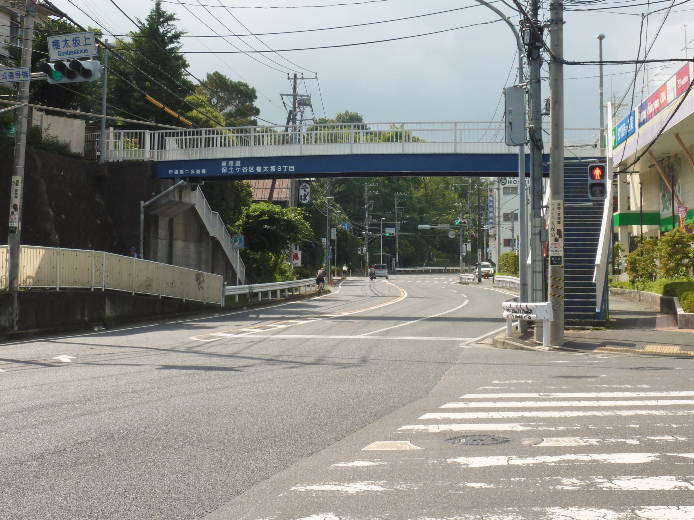 Stadtstraße mit einer Fußgängerbrücke darüber, Fahrzeuge auf der Straße, Strommasten mit Drähten, Verkehrsampeln, Schilder, Gebäude, Bäume, Pflanzen und einen Himmel als Hintergrund.
