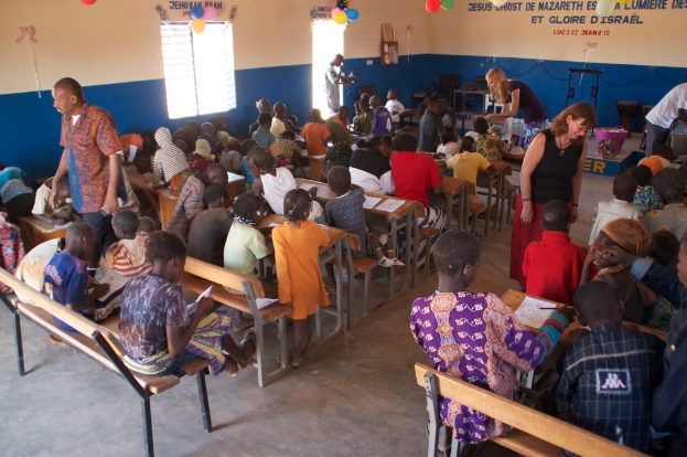 Kinder auf Bänken in Vordergrund mit Erwachsenen, die sie führen, Stühle, eine Tür, ein Fenster und ein Tisch im Hintergrund, Ballons und Text oben.