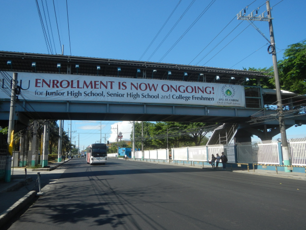 Ein Bus fährt unter einer Brücke mit einem "Jetzt ist Einschreibung möglich"-Schild, Menschen gehen auf dem Gehweg darunter, Strommasten mit Drähten, Bäume und ein klarer blauer Himmel im Hintergrund.