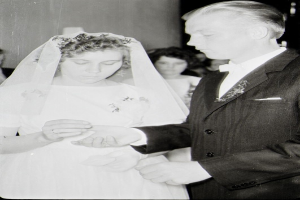 Ein Schwarz-Weiß-Foto einer Braut in einem weißen Kleid und Schleier und eines Bräutigams in einem Anzug, die sich ihre Eheringe austauschen, mit ein paar Menschen im Hintergrund.