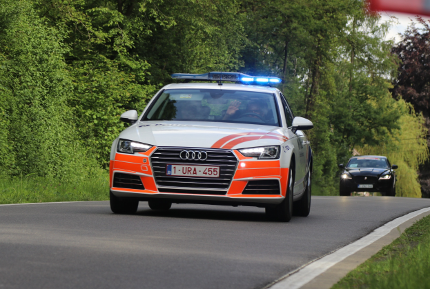 Ein Polizei-Audi A4 mit zwei Insassen fährt entlang einer grasbewachsenen Straße neben einem Wald unter einem klaren blauen Himmel.