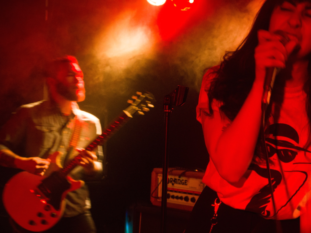 Eine Frau singt in ein Mikrofon auf der Bühne, wür ein Mann eine Akustikgitarre im Hintergrund spielt, mit Deckenbeleuchtung, die die Szenerie erhellt.