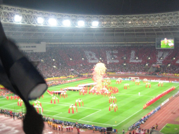 Ein Stadion mit Flutlicht, Eisengittern, Zuschauern, stehenden Personen, Werbetafeln, Anzeigetafeln, einer Überwachungskamera und Fahnen und Dekorationen.