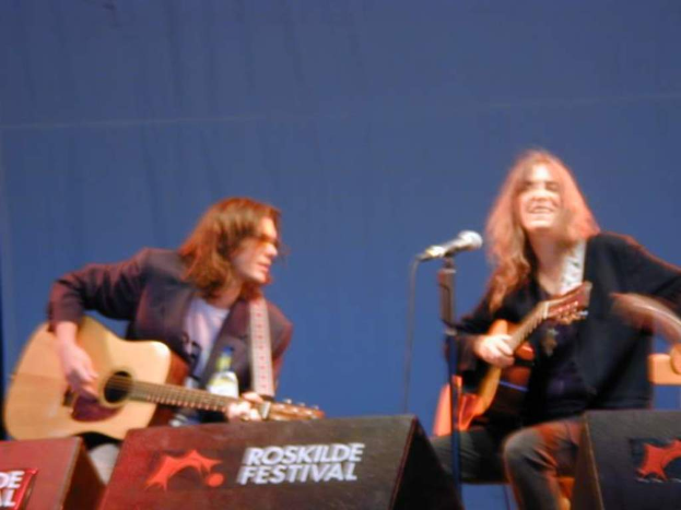 Zwei Männer auf der Bühne mit Musikinstrumenten, einem Mikrofon davor und zwei Kisten mit der Aufschrift "ROCK SLIDE FESTIVAL" davor.