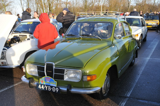 Mehrere Autos mit Menschen in der Nähe, die meisten tragen Jacken, vor dem Hintergrund von Bäumen, Pfählen und Himmel.