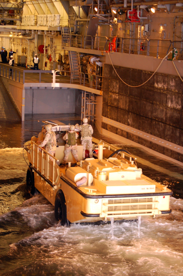 Amphibisches Fahrzeug fährt durch Wasser mit Helmträgern, neben einer Wand und Hintergrundelementen einschließlich Menschen, Geländern, Lampen und Rohren.