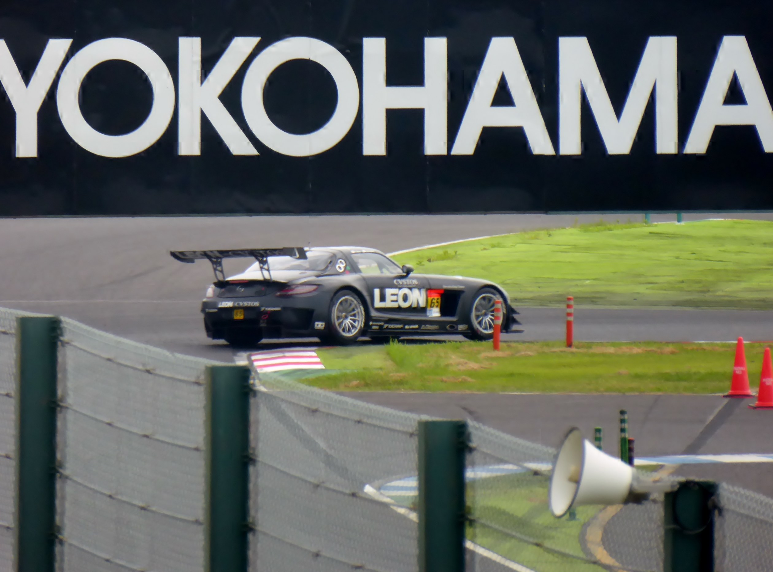 Schwarzer Sportwagen auf einer Rennstrecke mit Yokohama-Schriftzug, umgeben von Verkehrskegeln, Gras, Zaun und einem Lautsprecher im Vordergrund.