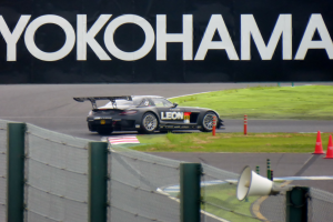 Schwarzer Sportwagen auf einer Rennstrecke mit Yokohama-Schriftzug, umgeben von Verkehrskegeln, Gras, Zaun und einem Lautsprecher im Vordergrund.