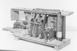 Ein altes Schwarz-Weiß-Foto einer großen, metallenen Druckerpresse mit Seitengriff und einem großen oberen Rad, das auf einem Holzpaletten gegen eine einfarbige Wand gestellt ist.