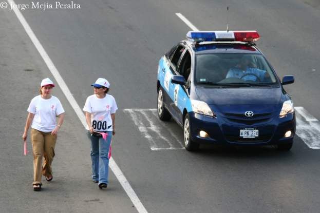 Zwei Frauen gehen auf einer Straße, eine hält einen rosa Zettel und trägt eine rosa und weiße Mütze, die andere hält einen rosa Zettel und trägt eine blaue und weiße Mütze, mit einem patrouillierenden Polizeiwagen und seinem Fahrer auf der rechten Seite.