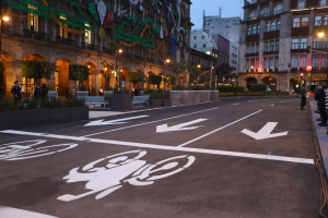 Eine Stadtstraße mit einem Fahrradweg, Gebäuden, Straßenlaternen, Verkehrspfosten, Ampeln, Menschen auf dem Gehweg, Bänken, Pflanzen, Bäumen, dekorativen Elementen und einem klaren blauen Himmel.