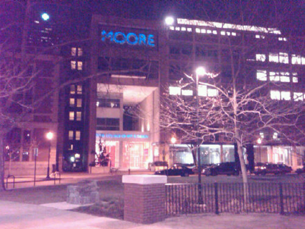 Ein Krankenhausgebäude mit mehreren Fahrzeugen und Bäumen davor, umgeben von einem Zaun.