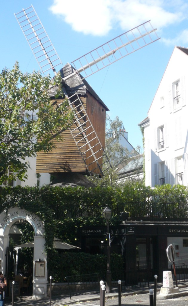 Ein Windrad, mehrere Gebäude, Bäume, Metallstangen und ein Zaun in einer ländlichen Umgebung.