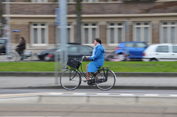 Zwei Personen fahren mit Fahrrädern auf einer Straße mit Gras, Pfosten, Autos und einem Gebäude im Hintergrund.