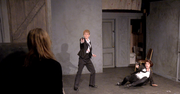 Zwei Frauen in schwarzen Anzügen, eine sitzend auf dem Boden und eine stehend, beide mit Waffen, sowie eine dritte Frau in der Nähe, in einem Raum mit einem Stuhl, einer Toilette und einer hölzernen Tür.