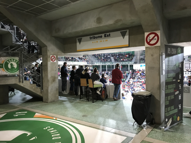 Eine Gruppe von Menschen steht vor einer sitzenden Menge in einem Stadion, mit Schildern, die "Boston Celtics vs New York Jets" und andere Stadion-Elemente anzeigen.