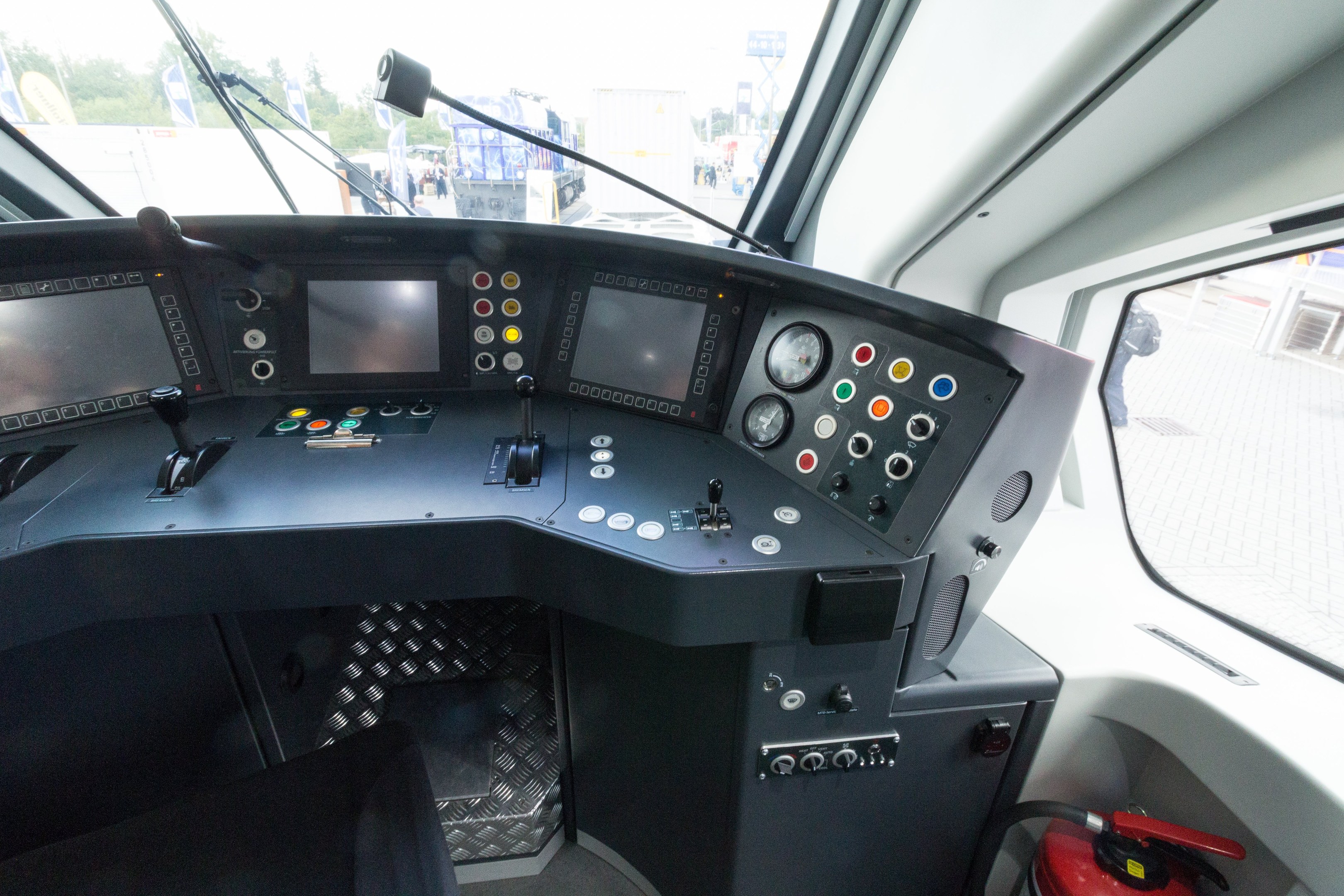 Cockpit eines Zuges mit einem Feuerlöscher im Vordergrund, das eine Glasscheibe zeigt, die Menschen, Fahrzeuge, Bäume und andere Gegenstände außerhalb zeigt.