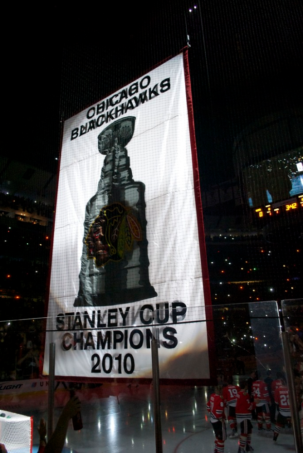 Eine nächtliche Stadionszene mit einem großen Banner in der Mitte, Zuschauern im Hintergrund, einem Glas auf dem Boden, einigen Spielern auf dem Feld und beleuchteten Tribünen.