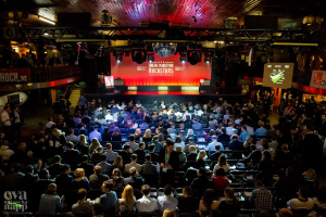 Eine große Menschenmenge sitzt auf Stühlen vor einer Bühne mit Bildschirmen und Plakaten, die von Scheinwerfern beleuchtet und von Geländern umgeben sind und gespannt auf den Beginn eines Online-Marketing-Rockstars-Events warten, mit einem Wasserzeichen in der linken unteren Ecke.