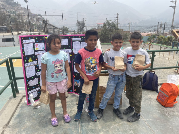 Gruppe von Kindern auf einem Basketballfeld mit Papiertüten stehend, mit Tüten und Gegenständen rechts und einer Tafel mit Text links, vor einem Hintergrund aus Zaun, Pfosten, Straßenlaternen, Bäumen, Gebäuden, Hügeln und Himmel.