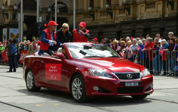 Ein Auto mit Menschen darin fährt auf einer Straße, mit verschiedenen Objekten, Menschen und Strukturen im Hintergrund.