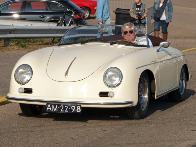 Ein Mann fährt einen weißen Porsche 356 Speedster auf einer Straße, umgeben von Menschen in verschiedenen farbigen Kleidern, mit einem Geländer und einem schwarzen Müllcontainer an der Seite und mehr Autos und einem Gebäude im Hintergrund.