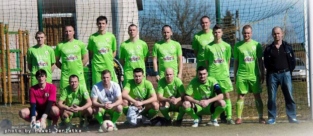 Eine Gruppe von Männern in grünen T-Shirts steht und hockt auf einem Fußballfeld mit einem Ball davor, ein Torpfosten rechts und ein Hintergrund aus Bäumen, Gebäuden, Pfosten, Fahrzeugen und dem Himmel.