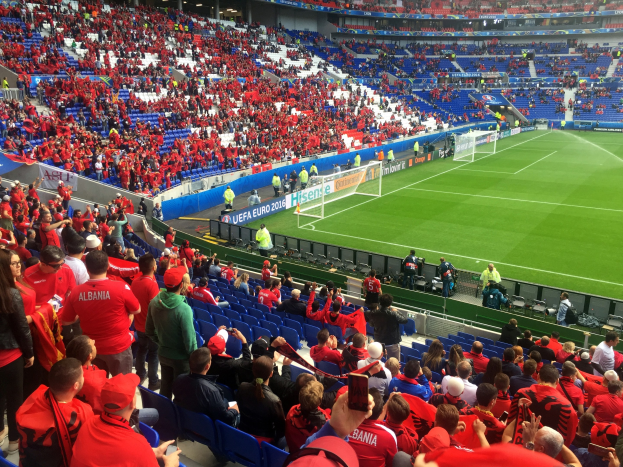 Eine große Menschenmenge in einem Stadion schaut einem Fußballspiel zu, mit Menschen, die auf Stühlen sitzen und stehen, Schildern, einem Torpfosten und Deckenleuchten.