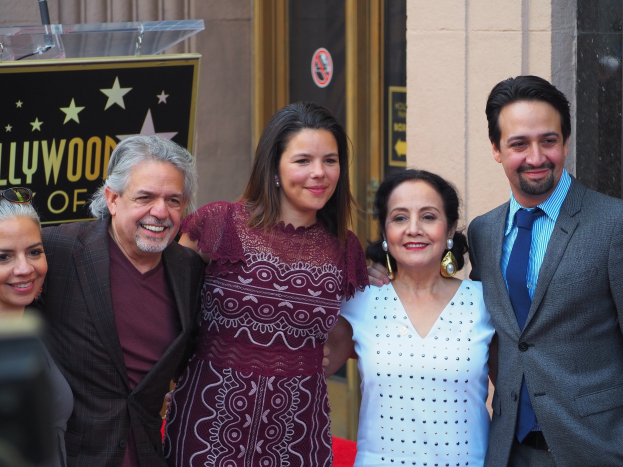 Eine Gruppe von Menschen, die auf einem roten Teppich stehen, lächeln und für ein Foto posieren, mit einer Kamera auf der linken Seite, einer Tafel mit Text, einem Glaspodium und einer Wand im Hintergrund.
