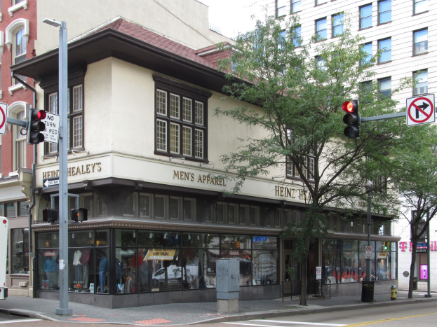 Eine belebte Straßenkreuzung mit einem geparkten Bus, Gebäuden, Bäumen, Laternen, Ampeln und Schildern, mit einem Kaufhaus im Hintergrund unter einem sichtbaren Himmel.