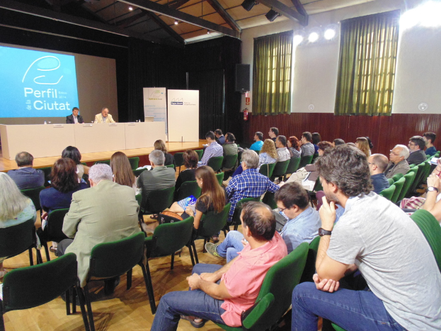 Eine Gruppe von Menschen sitzt auf grünen Stühlen in einem Raum mit Bannern, einem Tisch, einer Projektionswand, Vorhängen, Feuerlöschern und Lautsprecherboxen.