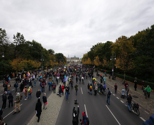 Eine große Gruppe von Menschen marschiert auf einer von Bäumen gesäumten Straße in Berlin, mit Kameras bewaffnet, vor einem Gebäude und einem klaren Himmel.