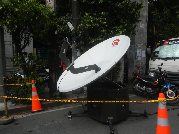 Eine Satellitenschüssel auf der Seite einer Straße, umgeben von Absperrgittern, Fahrzeugen, Menschen, Bäumen, Polen, Schildern und einem Gebäude im Hintergrund.