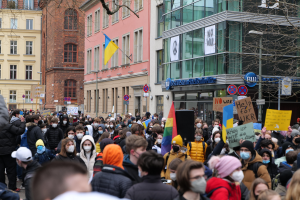 Eine große Gruppe von maskierten Menschen mit Schildern protestiert gegen eine Regierungsentscheidung, Masken auf der Straße zu verbieten, umgeben von Gebäuden, Schildern, Fahnen und Bäumen.