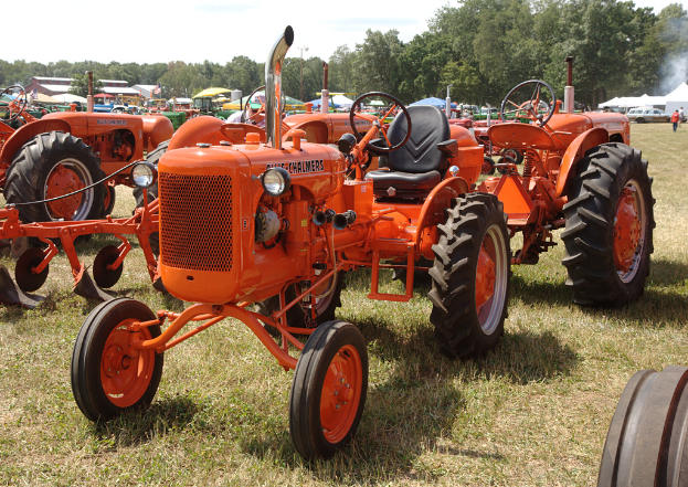 Orangene Traktoren auf einem Rasen mit Gras unten, verschiedene Fahrzeuge, grüne, blaue und weiße Schirmzelte, Bäume, Gebäude und zwei Männer rechts stehend, unter einem Himmel oben.