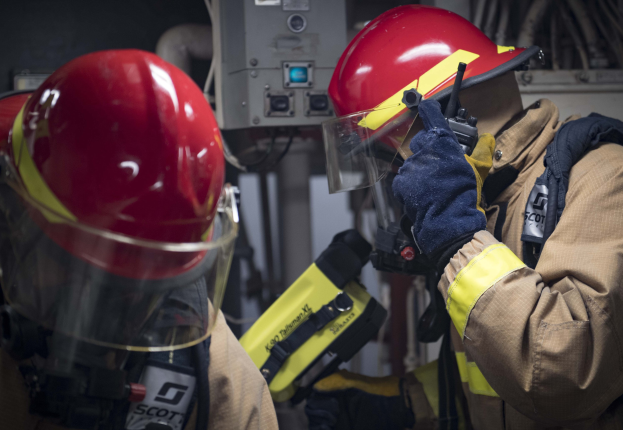 Zwei Feuerwehrleute in Schutzausrüstung bei der Arbeit an einem Feuerhydranten während einer übung mit Maschinen und Kabeln im Hintergrund.