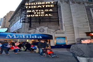Eine Gruppe von Menschen, die auf einer Straße mit Gebäuden und Werbetafeln im Hintergrund gehen, mit einem Baby in einem Kinderwagen darunter.